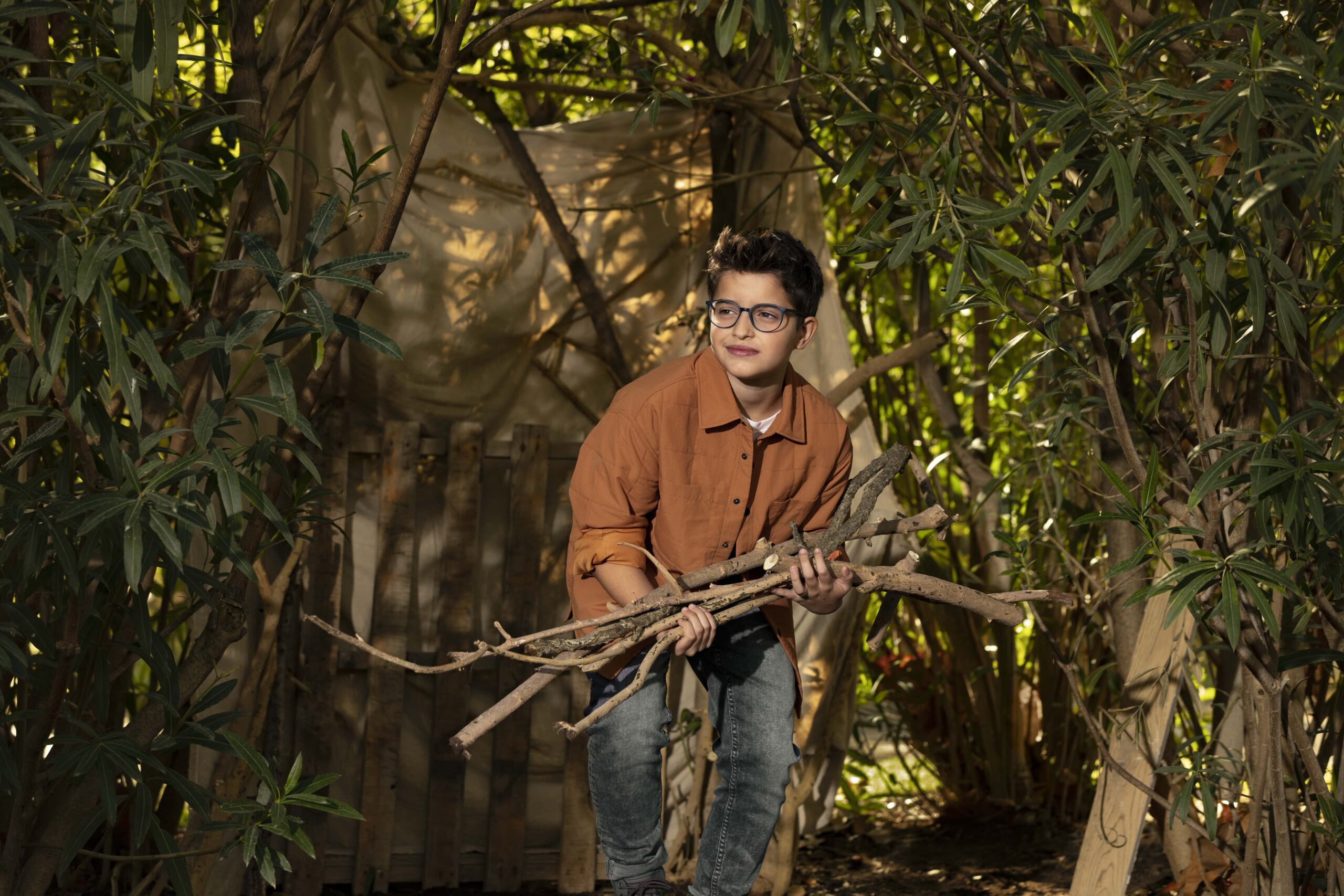 Children's Glasses , Children's Eye Tests A boy in a woodland, building a den. He has light brown hair. He wear a pair of HOYA MiYoSmart glasses, a burnt orange jacket and a pair of jeans. He's carrying a bunch of large sticks to build his den.