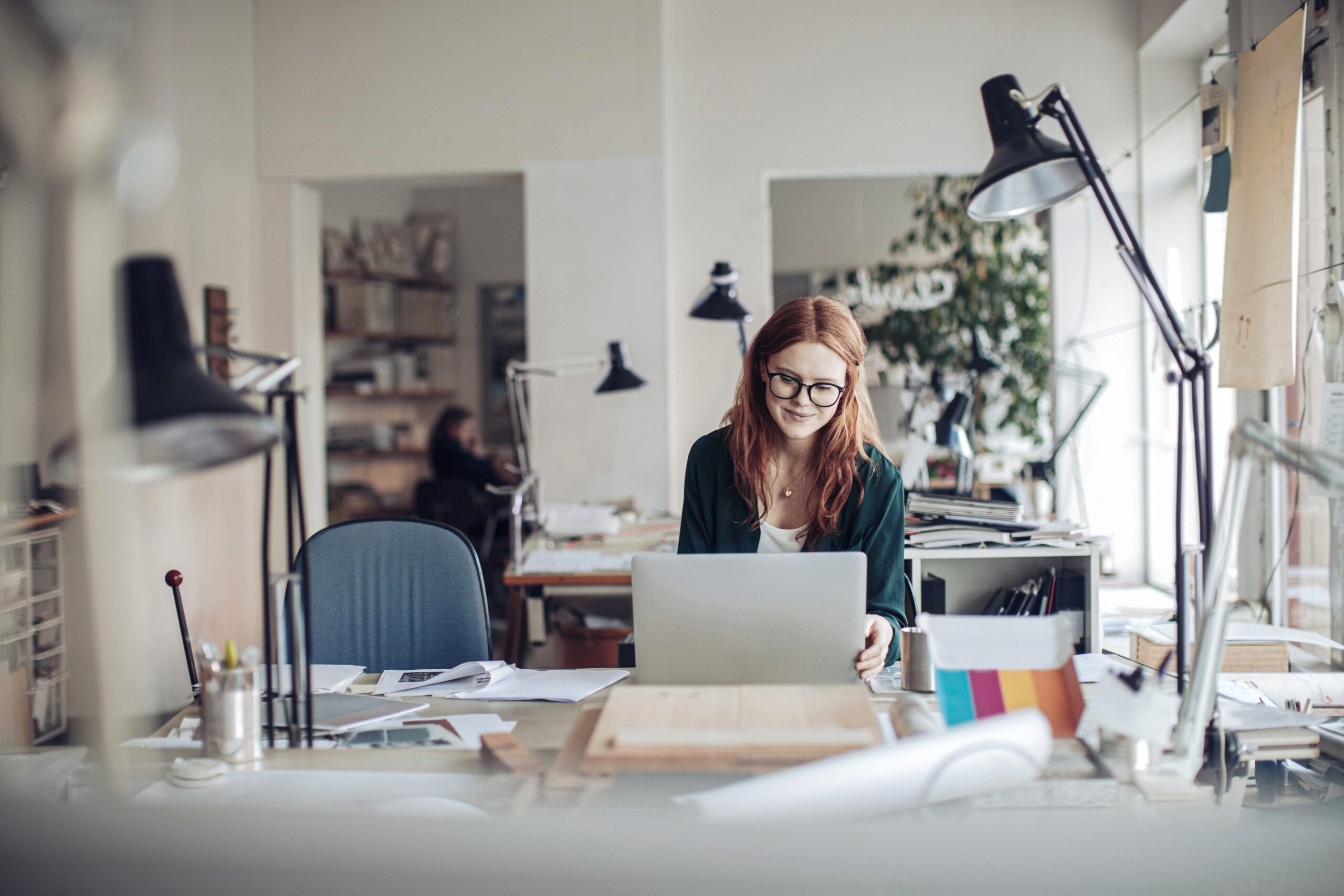 A young woman with red hair and glasses works at a desk in a bright, open studio space, looking at her laptop with a smile. The workspace is filled with various office supplies, papers, and desk lamps, creating a creative, organized environment. Other desks and a colleague are visible in the background. Artificial Light and Eye Health.