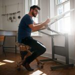 A man wearing glasses sits at a drafting table in a bright room, drawing or writing with a pen. Sunlight streams through the windows, illuminating his workspace. The room features wooden floors, simple furnishings, and art supplies hanging on the wall, giving it a creative, minimalist atmosphere.