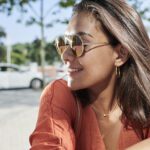 A woman smiling outdoors on a sunny day, wearing round, light-tinted sunglasses and a burnt orange top. She has shoulder-length brown hair and is accessorized with gold hoop earrings and a delicate necklace. Trees and a parked car are visible in the blurred background.