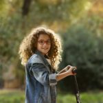 Girl with brown curly hair, wearing red glasses and a denim jacket is looking at the camera whilst she's riding her scooter through a park