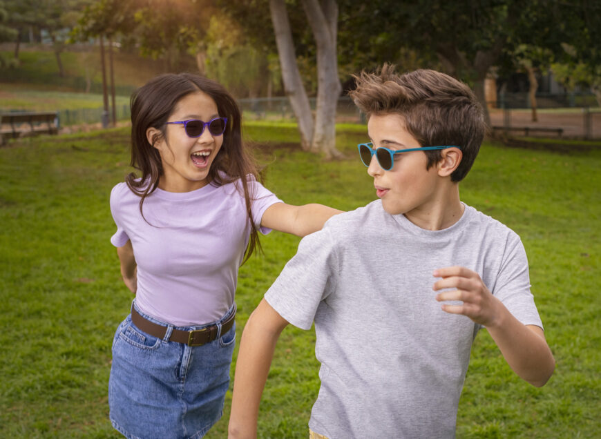 A preteen girl and a preteen boy in a parkland, playing tig. The girl is chasing the boy and looks like she's about to catch him. The girl has long dark brown hair, is wearing a lilac t shirt and a blue denim skirt and belt. She's wearing a pair of purple HOYA MiYoSmart Sunglasses. The boy has light brown hair and is wearing a marl grey t shirt. He's wearing a pair of turquoise HOYA MiYoSmart Lenses.