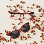 A pair of red glasses with accompanying sunglasses clip on, on top of castor seeds, out of which the oil is extracted and turned in to bio-based plastics, making sustainable eco-friendly glasses.