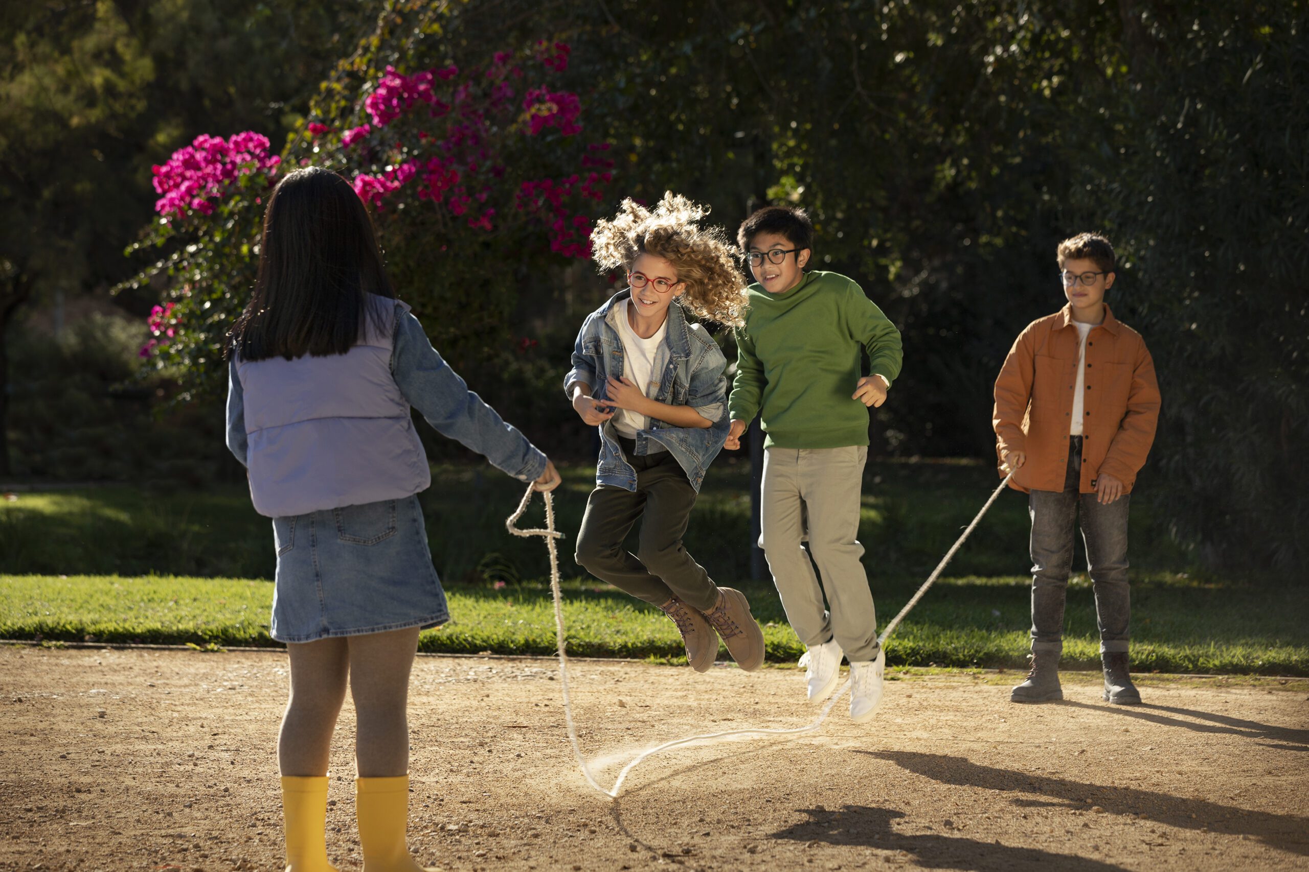 An image of four children playing with a skipping rope. The floor is beige coloured stone where the children are skipping. Behind that there is grass with lots of green foliage and bright pink flowers in one of the trees. Closest to the camera is a girl with her back to the camera, She is holding one end of the skipping rope. She has long black hair. She is wearing a blue denim long sleeve top and skirt, with a lilac bodywarmer, beige tights and yellow wellies. The next nearest to the camera is a girl who is smiling and jumping over the skipping rope. She is facing the girl holding the skipping rope. This girl has dark blonde, curly hair. She wears a pair of red HOYA MiYoSmart Glasses, a blue denim jacket, a white t shirt, dark green trousers and beige lace up boots. Behind her is a boy who is also smiling while jumping over the skipping rope. He has black short hair. He is wearing black round HOYA MiYoSmart Glasses, a grass green hoodie, light beige trousers and white trainers. Behind him is a boy who is holding the other end of the skipping rope. He has brown short hair. He wears black HOYA MiYoSmart Glasses, a burnt orange jacket, with a white t shite underneath, dark grey trousers and black lace up boots.