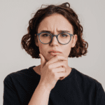 Young woman with short brown curly hair, wearing grey round glasses and a black t shirt looking quizzical.